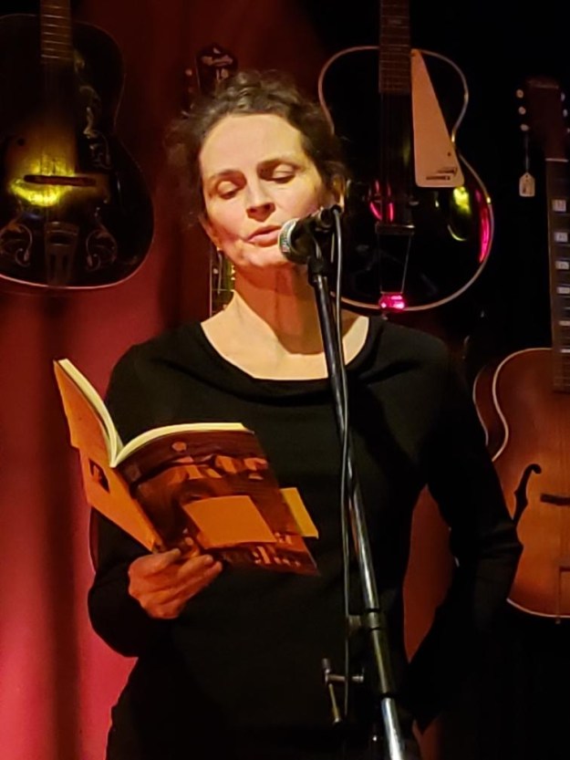 Jessice Cuello reads from Liar at a microphone. The room is dim and there are guitars hanging from the wall behind her.